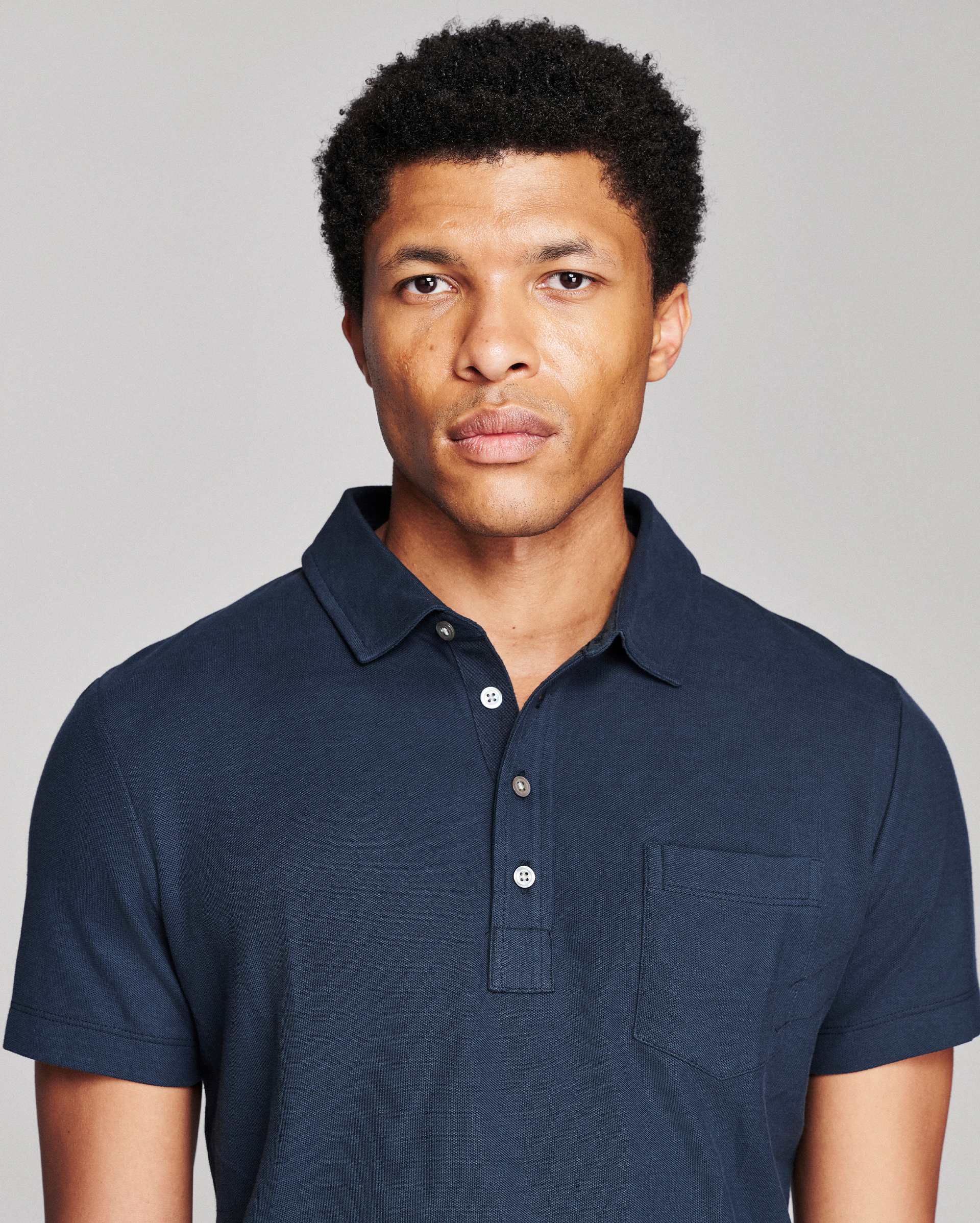 A man wearing a dark navy blue short-sleeve polo shirt with a three-button placket and a chest pocket, photographed against a solid light gray background.