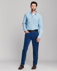 A man modeling a light blue long-sleeve button-up shirt and dark blue jeans with a black belt, standing against a neutral gray studio background.