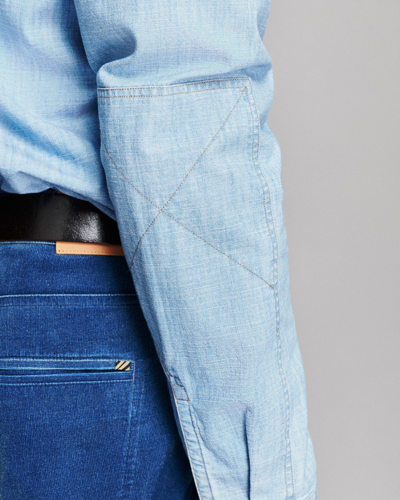 Close-up of a person wearing a light blue chambray long-sleeve shirt tucked into dark blue jeans with a black belt, showing the shirt's pocket and fabric texture.