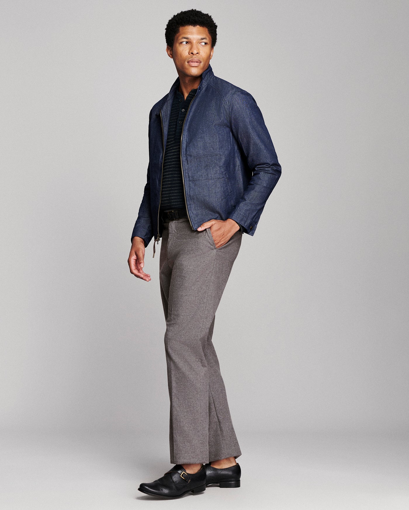 A man modeling a navy blue denim jacket over a black striped shirt, paired with grey flared trousers and black leather shoes, against a light gray studio background.