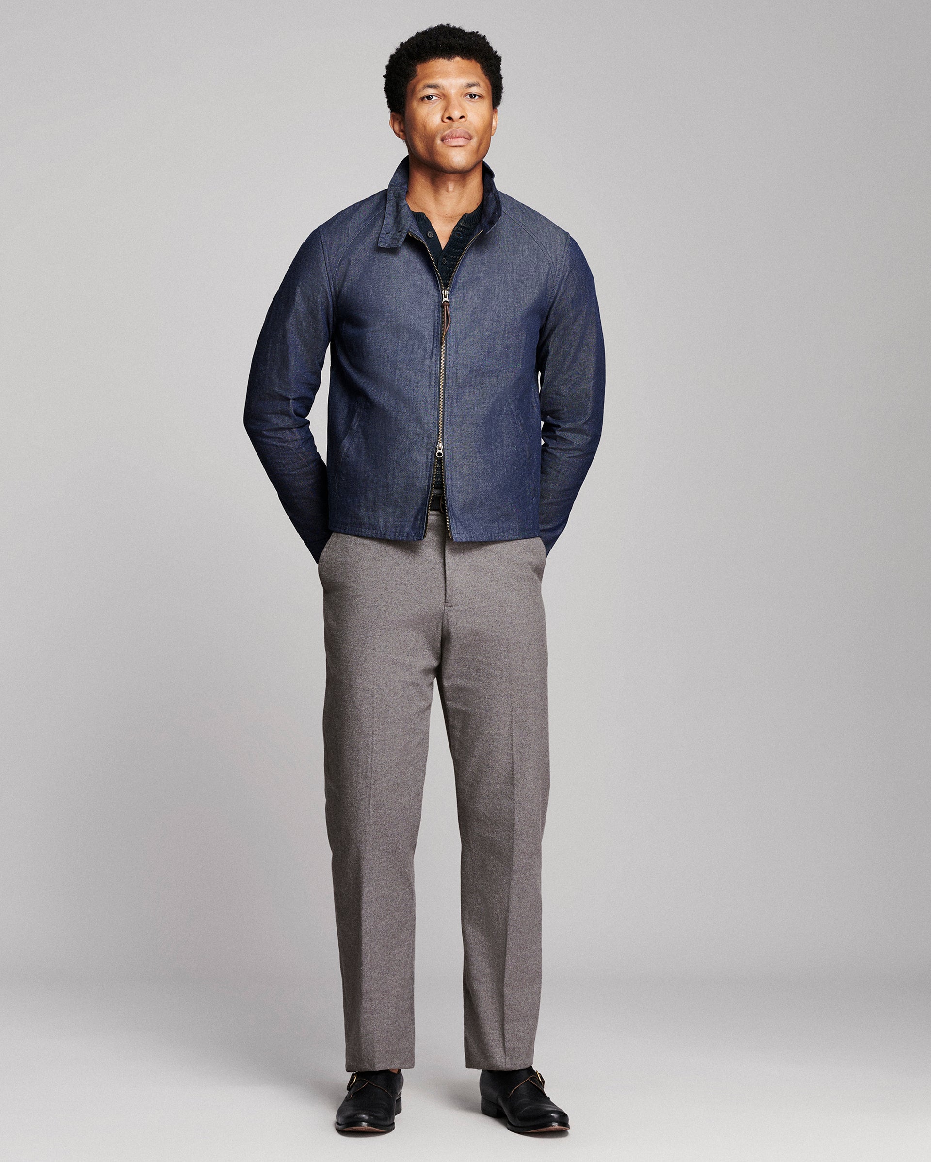 A man modeling a dark blue zip-up jacket with a dark collar, paired with gray trousers and black shoes, standing against a light gray studio background.