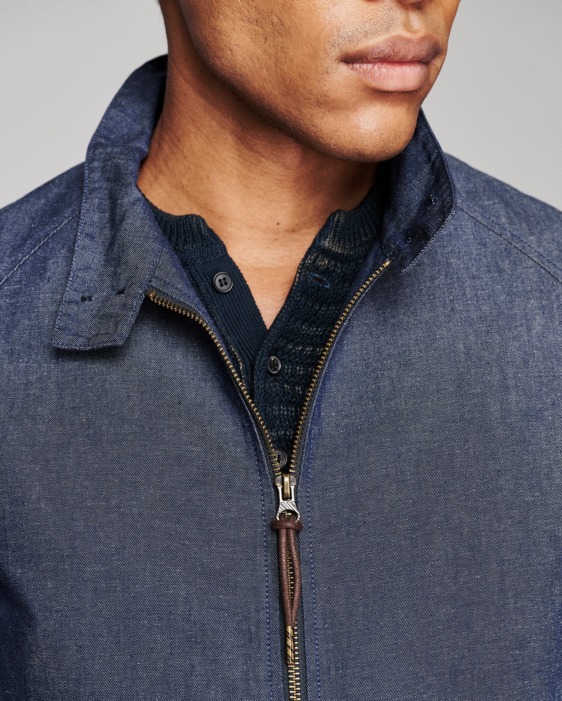 Close-up of a man wearing a blue textured zip-up jacket over a dark navy knit shirt, showing the collar, zipper, and fabric details against a neutral gray background.