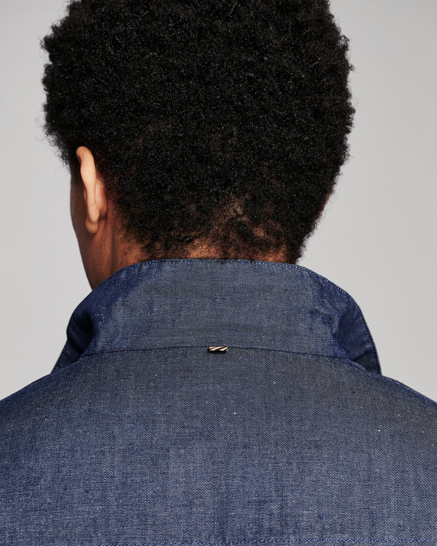 Back view of a person with short dark curly hair wearing a textured blue denim or chambray collared shirt jacket against a plain light gray background.