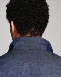 Back view of a person with short dark curly hair wearing a textured blue denim or chambray collared shirt jacket against a plain light gray background.