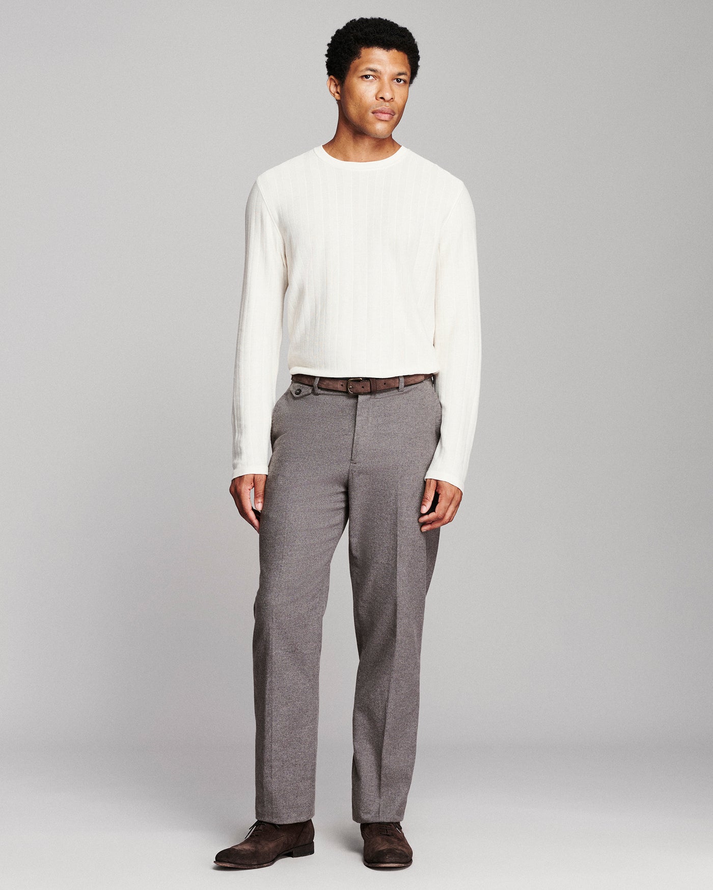 A man modeling a white ribbed long-sleeve knit sweater and gray pleated trousers with a brown belt and dark brown shoes, standing against a light gray studio background.