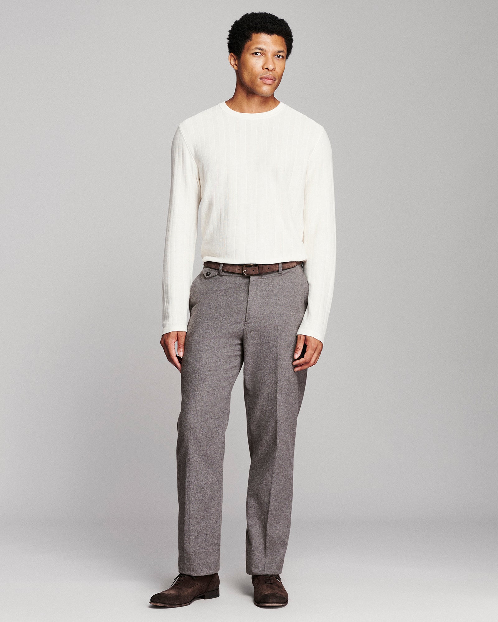 A man modeling a white ribbed long-sleeve knit sweater and gray pleated trousers with a brown belt and dark brown shoes, standing against a light gray studio background.
