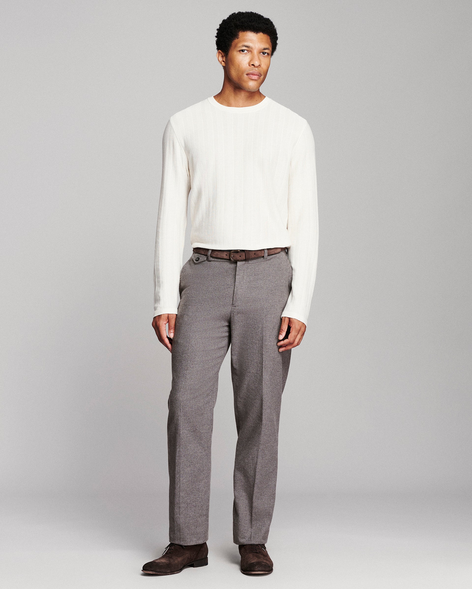 A man modeling a white ribbed long-sleeve knit sweater and gray pleated trousers with a brown belt and dark brown shoes, standing against a light gray studio background.