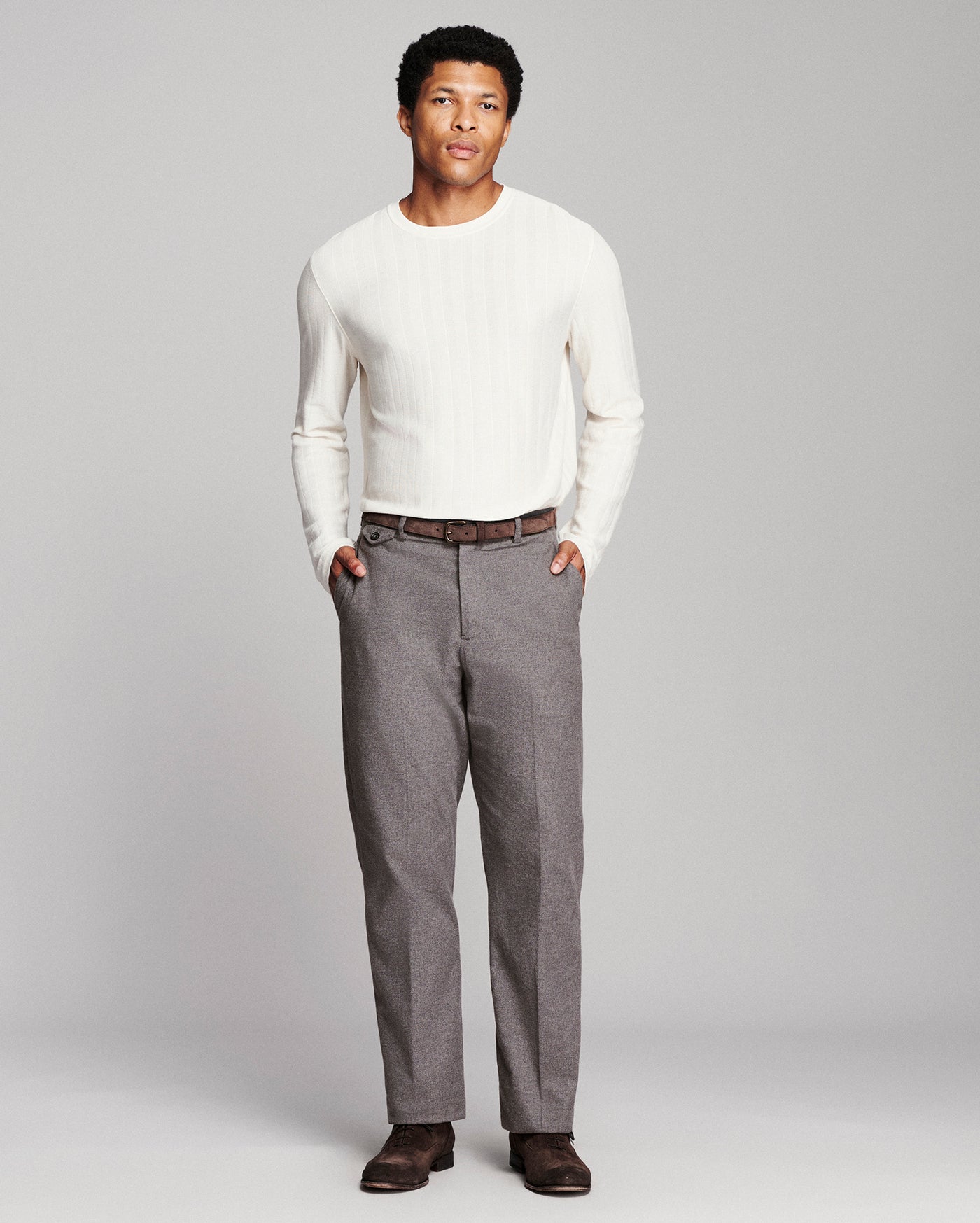 A man modeling a white ribbed long-sleeve knit sweater and gray trousers against a light gray studio background.