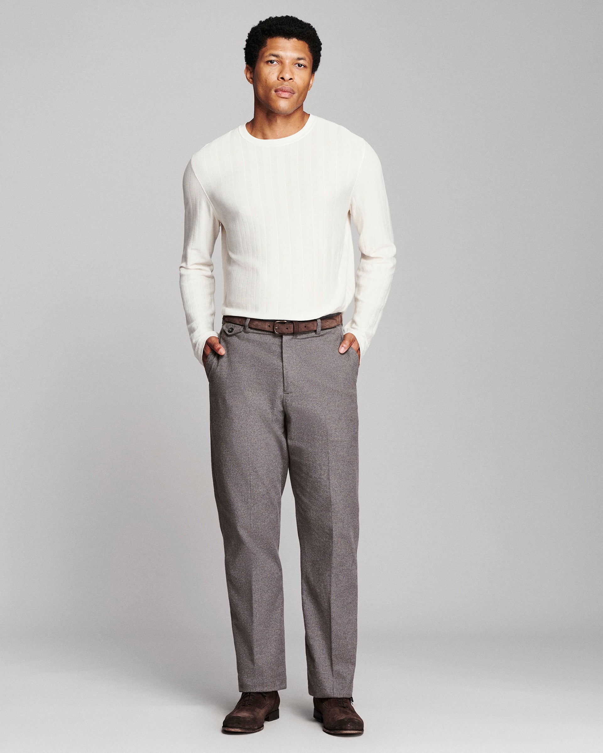 A man modeling a white ribbed long-sleeve knit sweater and gray trousers against a light gray studio background.