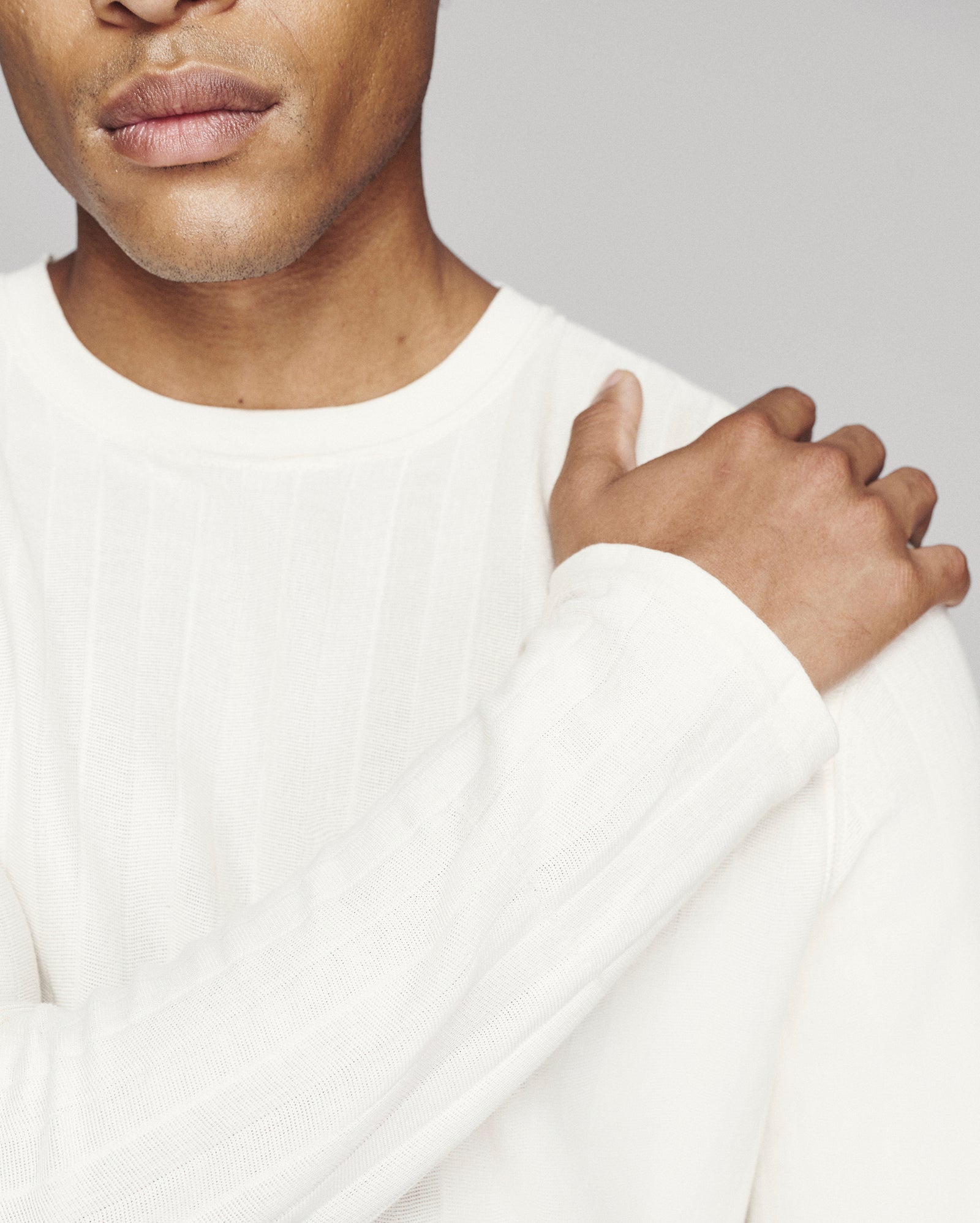 Close-up of a man wearing a white ribbed long-sleeve crewneck sweater, with his hand resting on his shoulder against a light gray background.