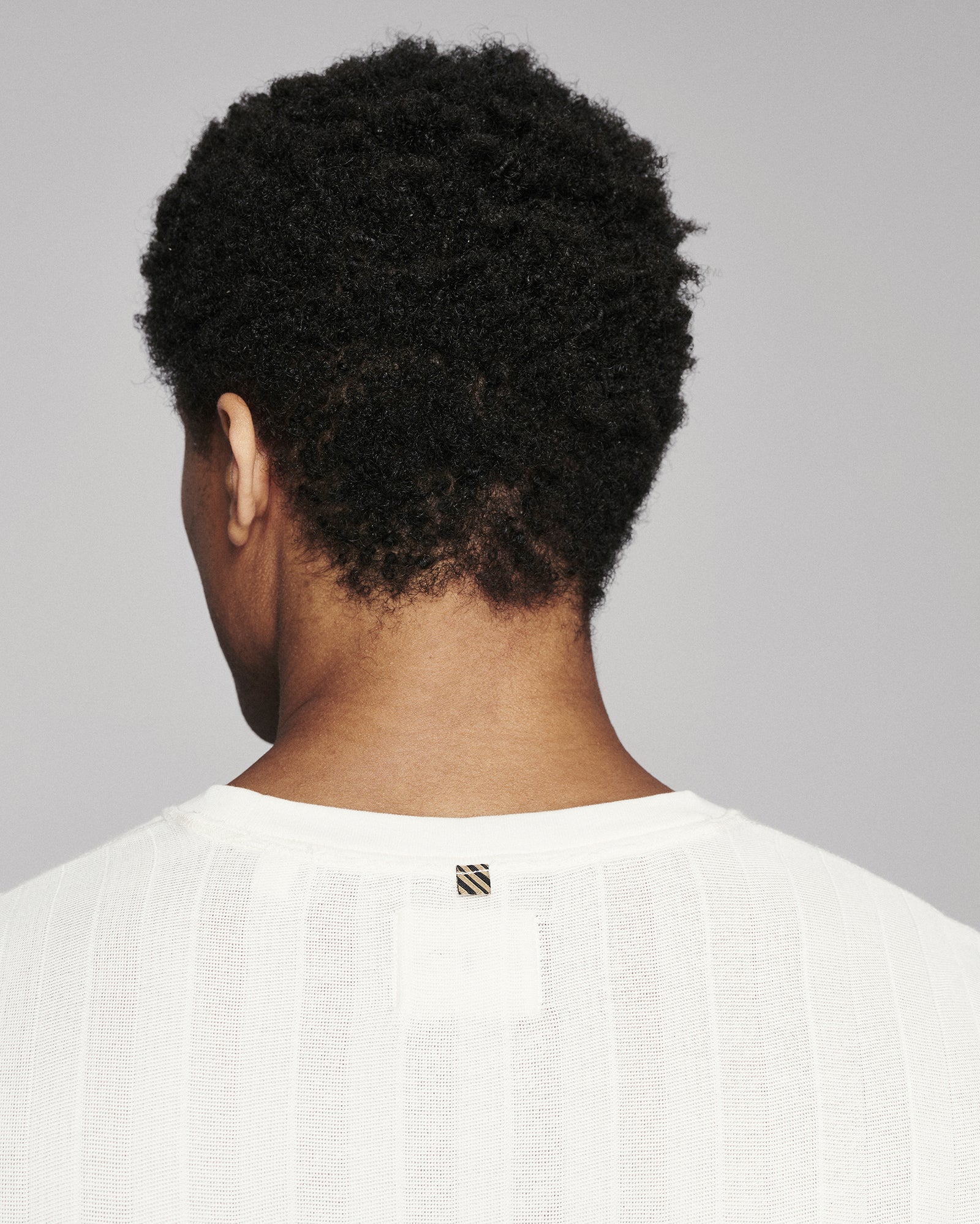 Back view of a person with short, curly black hair wearing a white textured knit top against a light gray background.