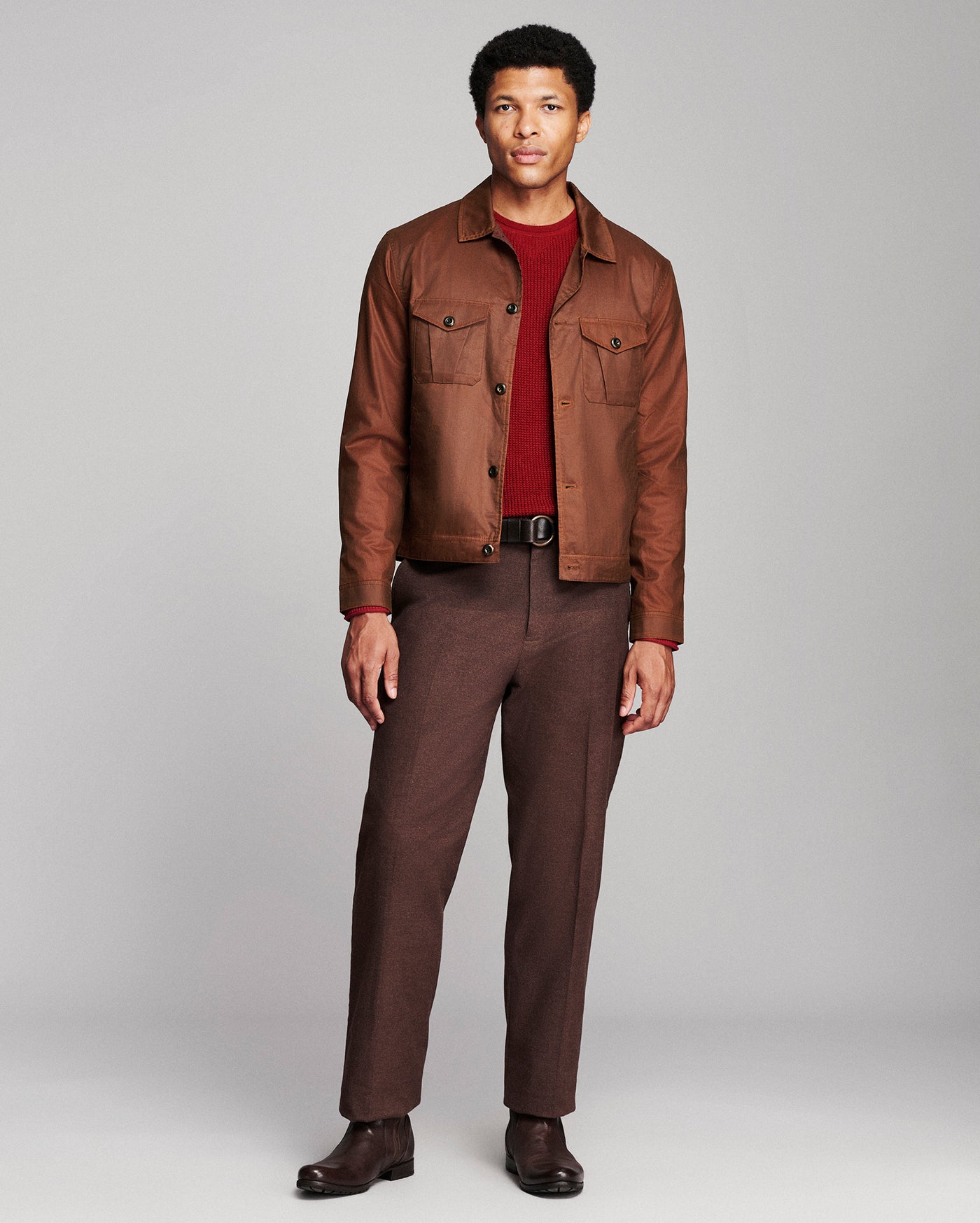A man modeling a brown leather jacket over a dark red t-shirt, paired with brown trousers and dark brown shoes, against a light gray studio background.