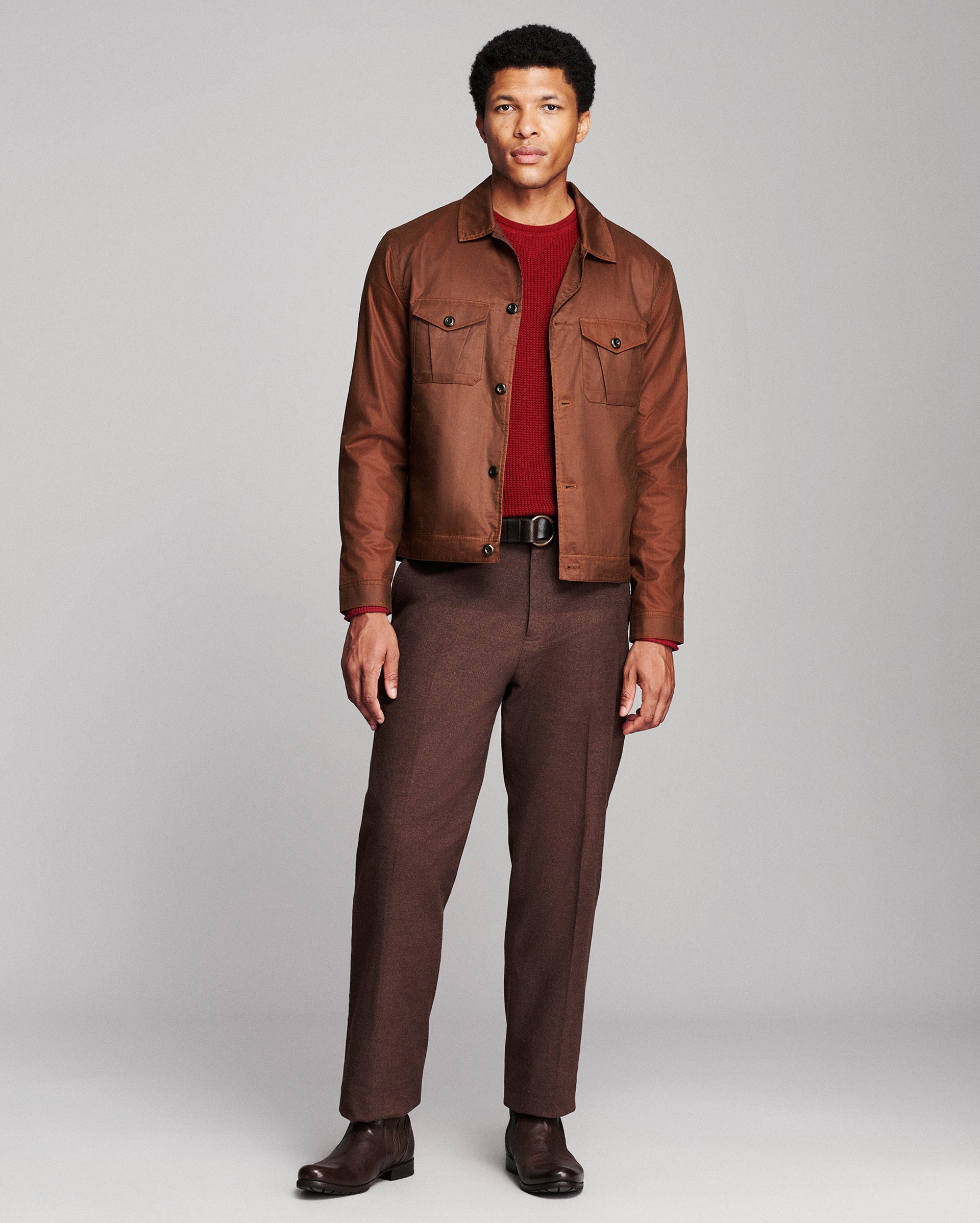 A man modeling a brown leather jacket over a dark red t-shirt, paired with brown trousers and dark brown shoes, against a light gray studio background.