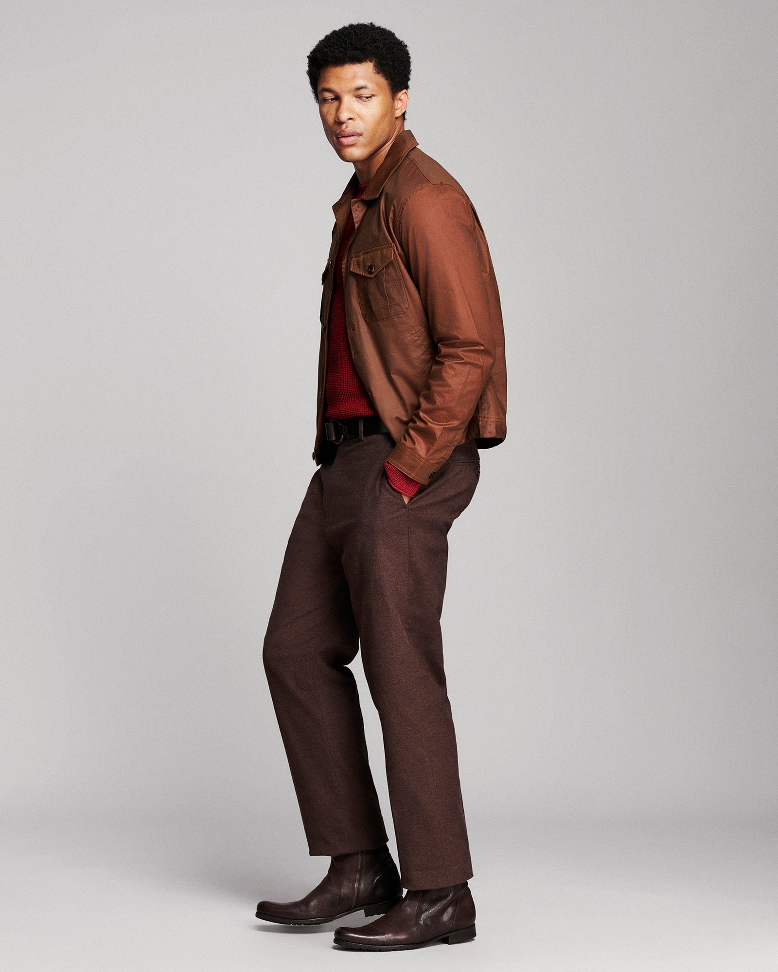 A man modeling a brown leather jacket over a dark red shirt, paired with dark brown trousers and boots, standing against a light gray studio background.