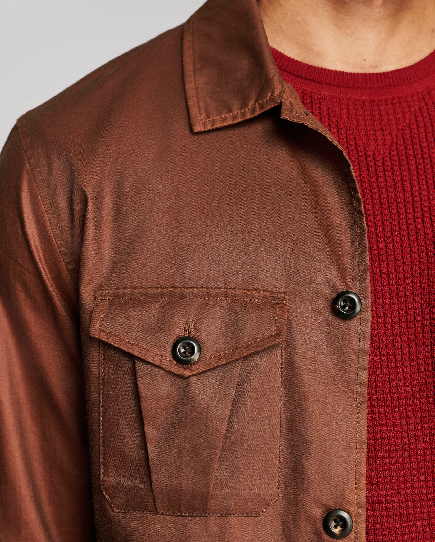 Close-up of a brown waxed cotton overshirt jacket worn over a red knit sweater, showing the chest pocket and buttons.