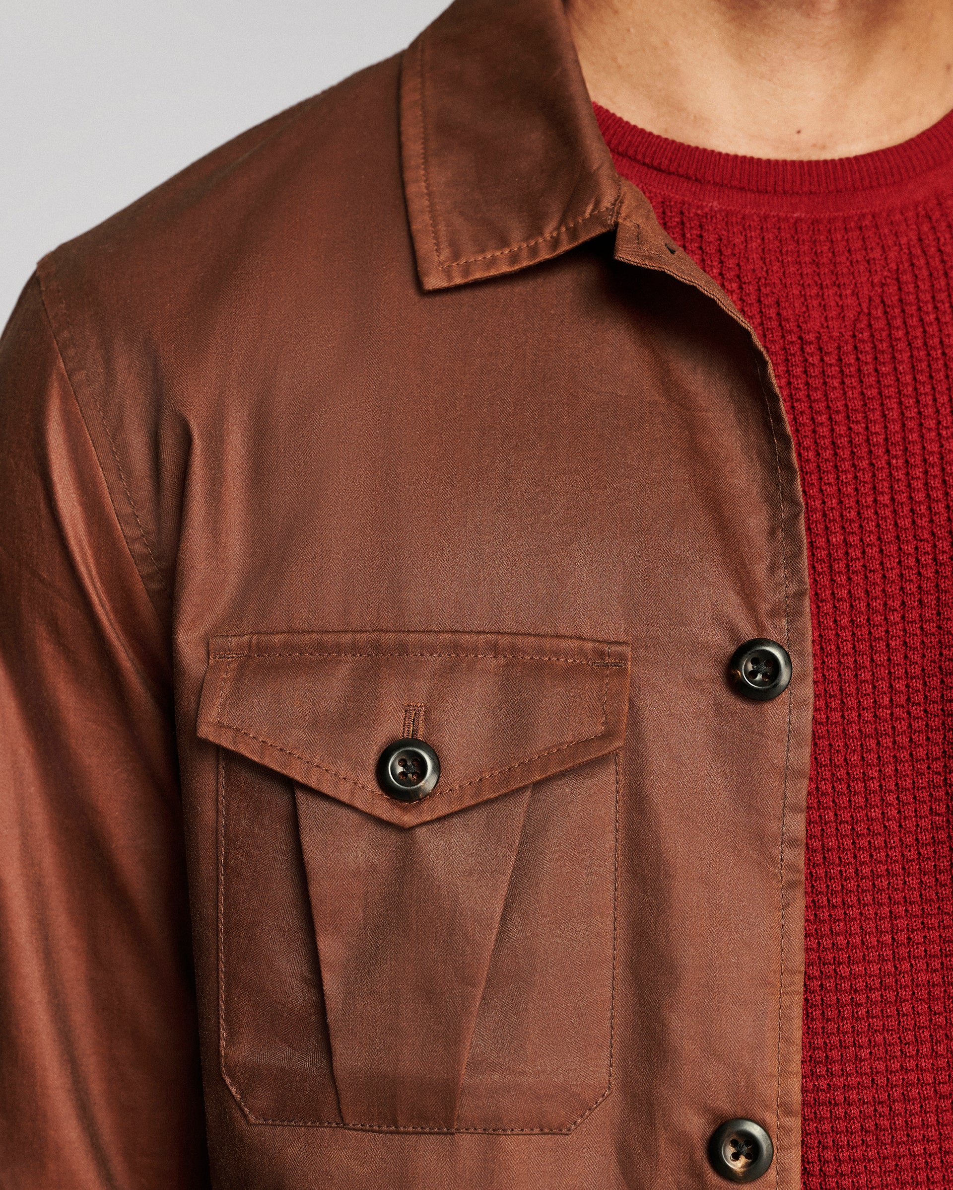 Close-up of a brown waxed cotton overshirt jacket worn over a red knit sweater, showing the chest pocket and buttons.