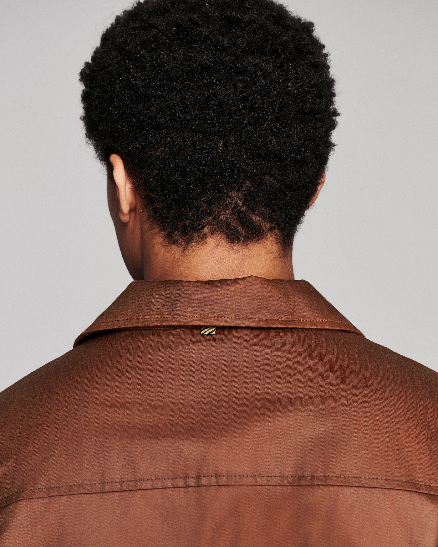 Back view of a person wearing a brown collared jacket against a light gray background.