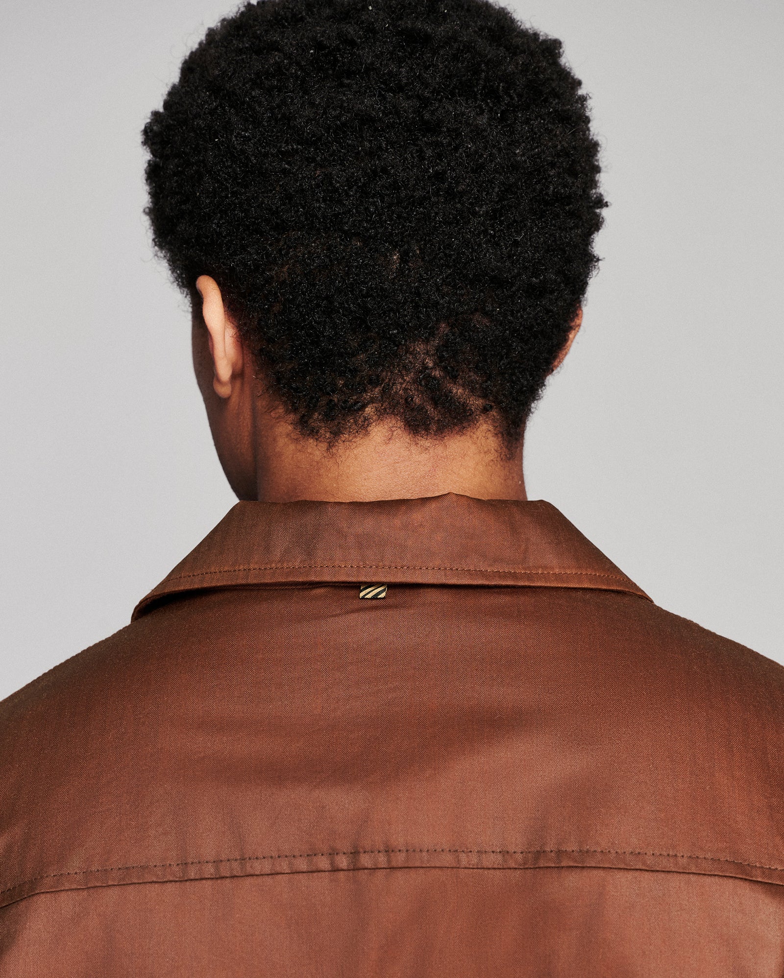 Back view of a person wearing a brown collared jacket against a light gray background.