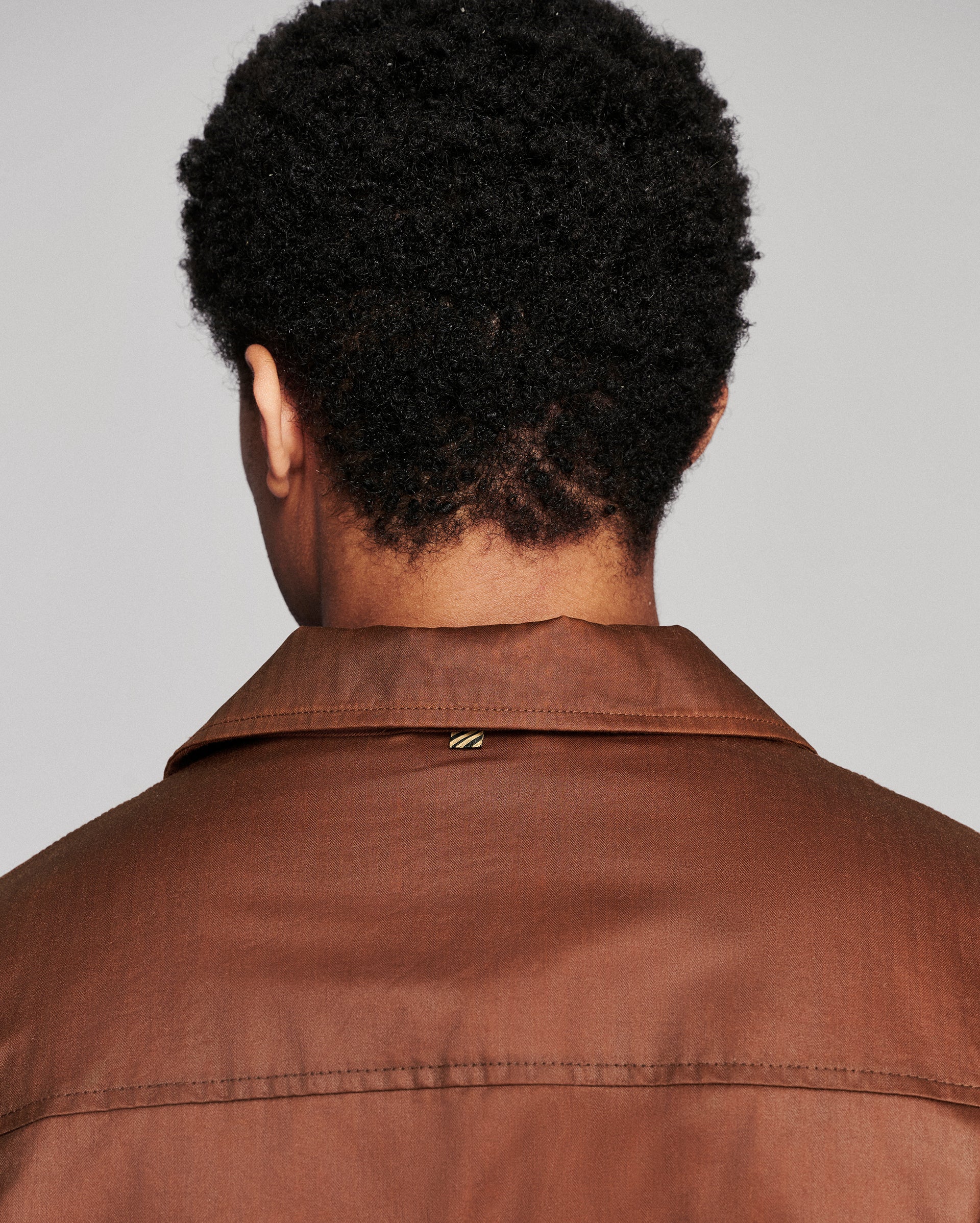 Back view of a person wearing a brown collared jacket against a light gray background.