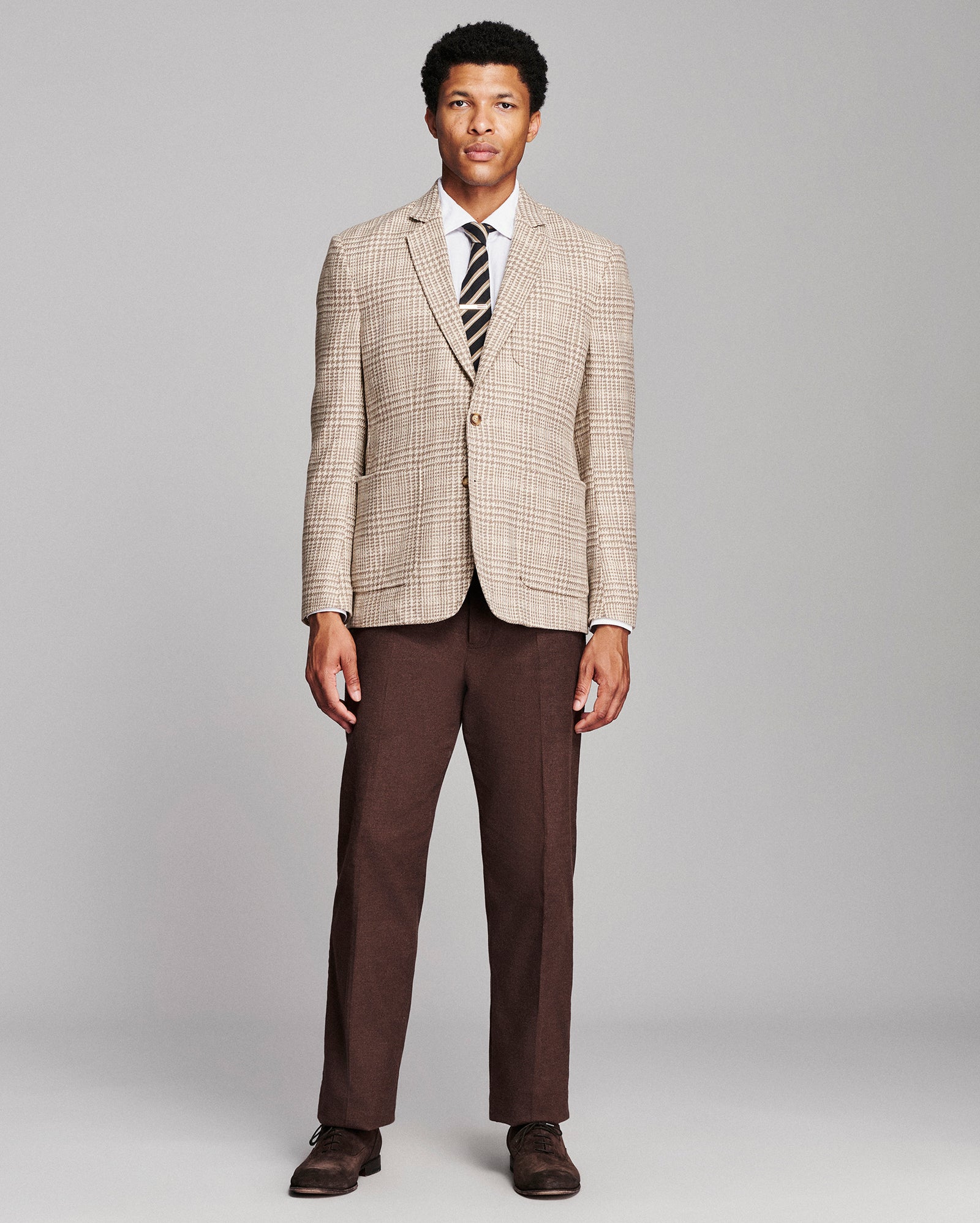 A man modeling a beige and brown plaid blazer with dark brown trousers, a white dress shirt, a striped tie, and brown dress shoes, standing against a light gray studio background.