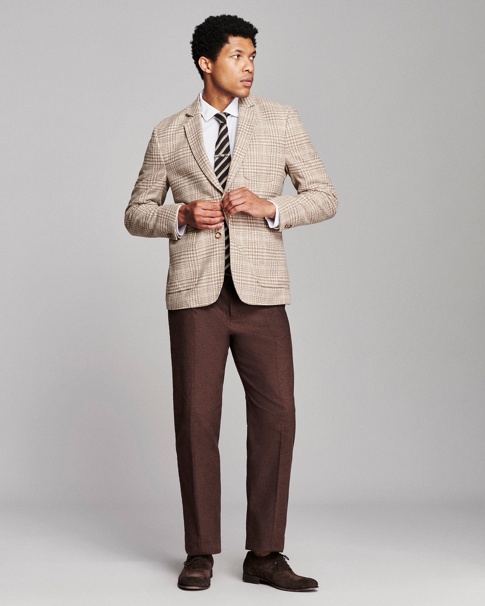 A man modeling a beige and brown checkered blazer, brown trousers, a white dress shirt, a striped tie, and brown dress shoes against a solid gray studio background.