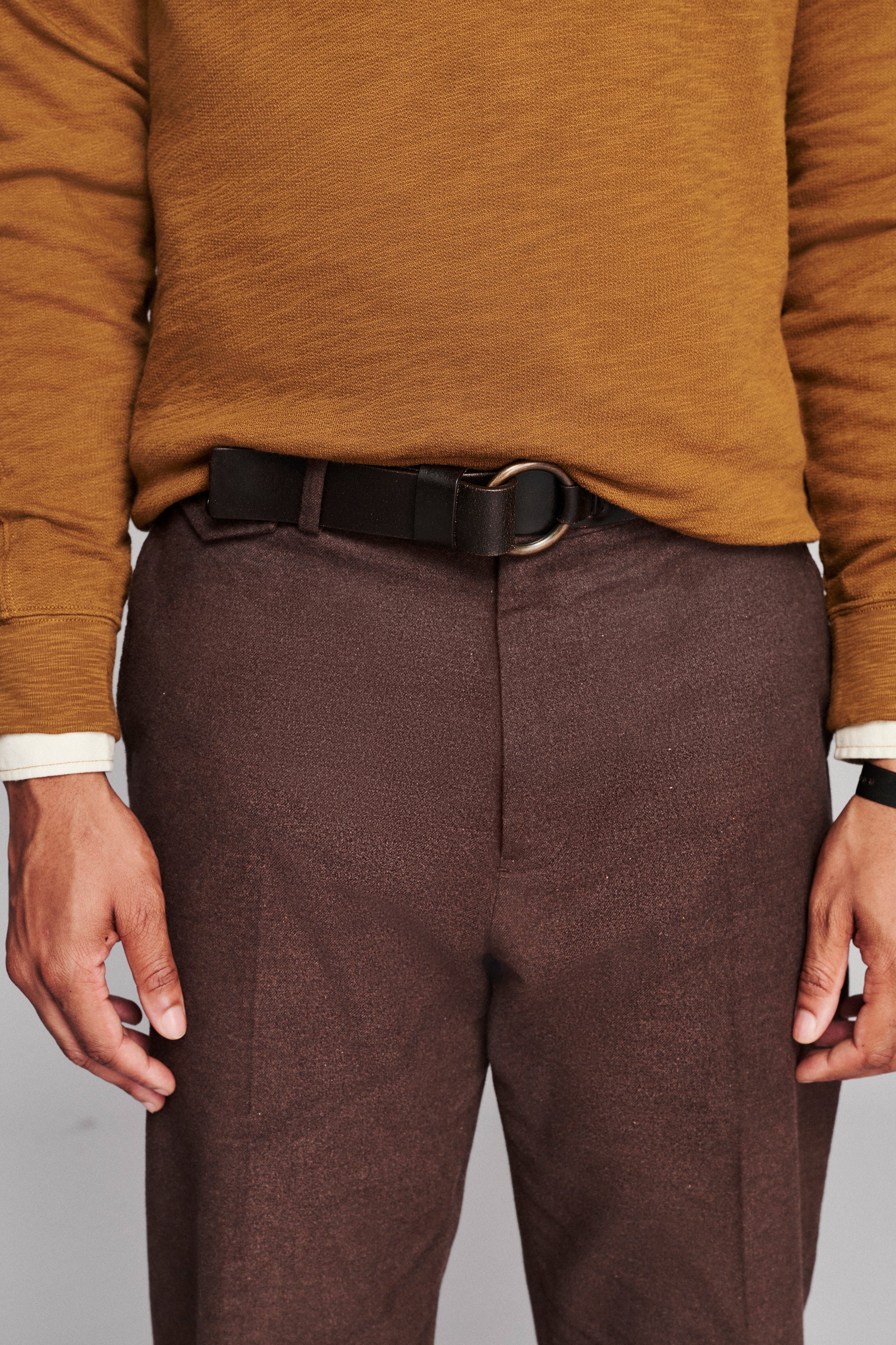 A close-up of a person wearing textured brown trousers, a black leather belt with a silver-tone oval buckle, and a mustard yellow knit sweater.
