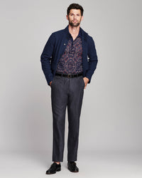 A man modeling a navy blue jacket over a patterned button-down shirt, paired with dark gray trousers, a black belt, and black leather dress shoes, against a light gray studio background.