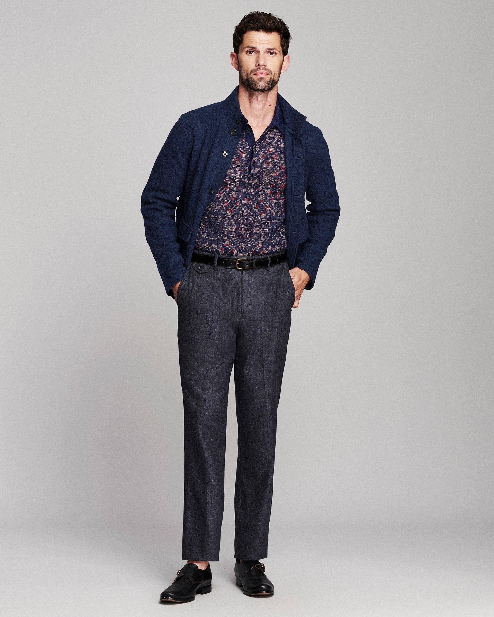 A man modeling a navy blue jacket over a patterned button-down shirt, paired with dark gray trousers, a black belt, and black leather dress shoes, against a light gray studio background.