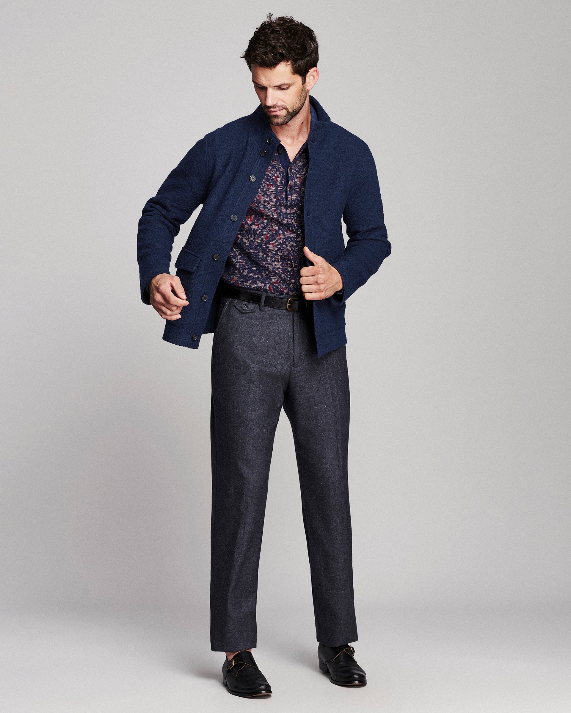 A man modeling a navy blue knit cardigan over a patterned shirt, paired with dark charcoal trousers and black loafers, against a light gray studio background.