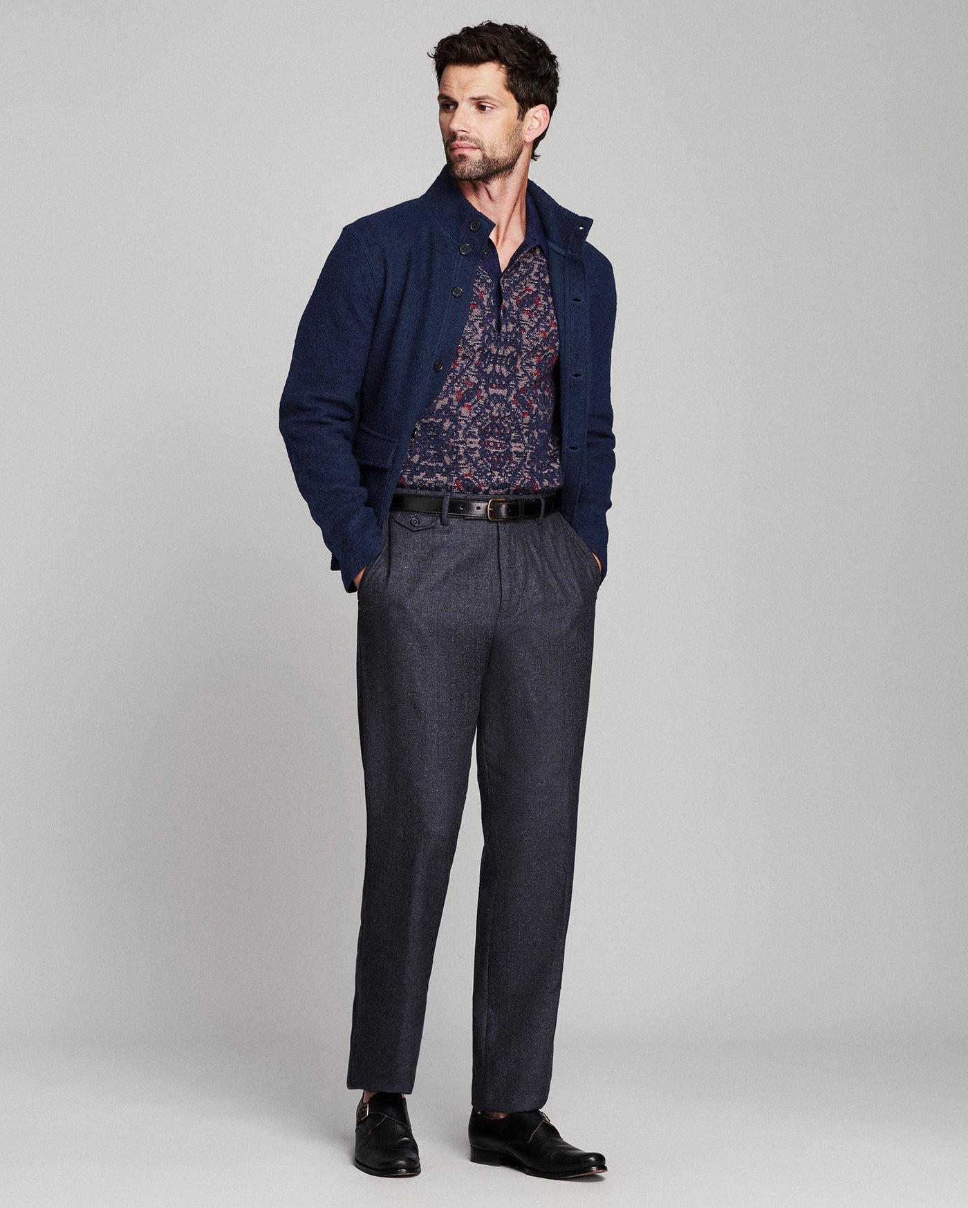 A man modeling a navy blue knit cardigan over a patterned shirt, paired with dark gray trousers, a black belt, and black dress shoes, against a light gray studio background.