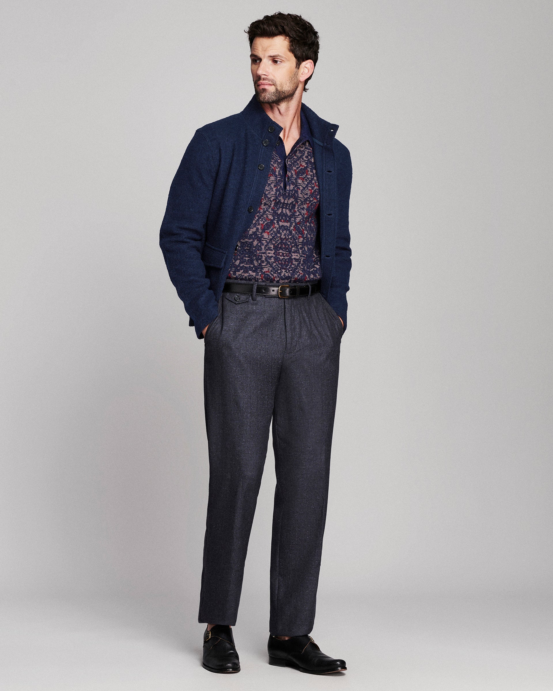 A man modeling a navy blue knit cardigan over a patterned shirt, paired with dark gray trousers, a black belt, and black dress shoes, against a light gray studio background.