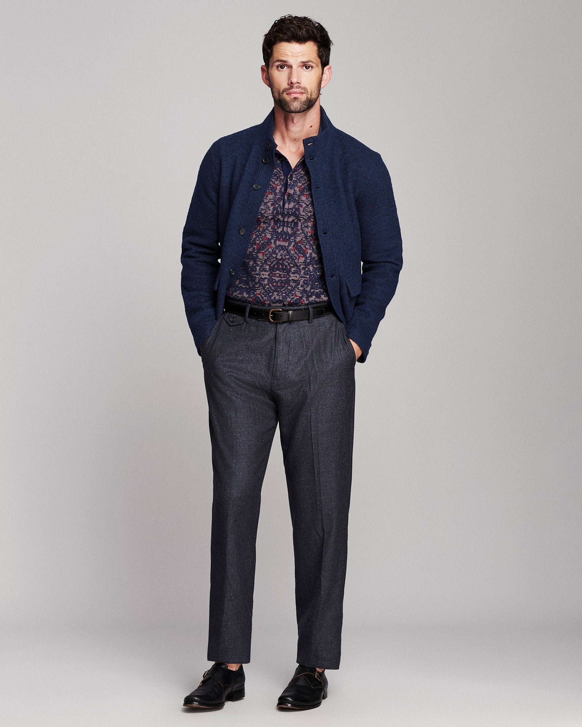 A man modeling a dark navy blue knit cardigan over a patterned button-down shirt, paired with dark gray trousers and black dress shoes, against a light gray studio background.