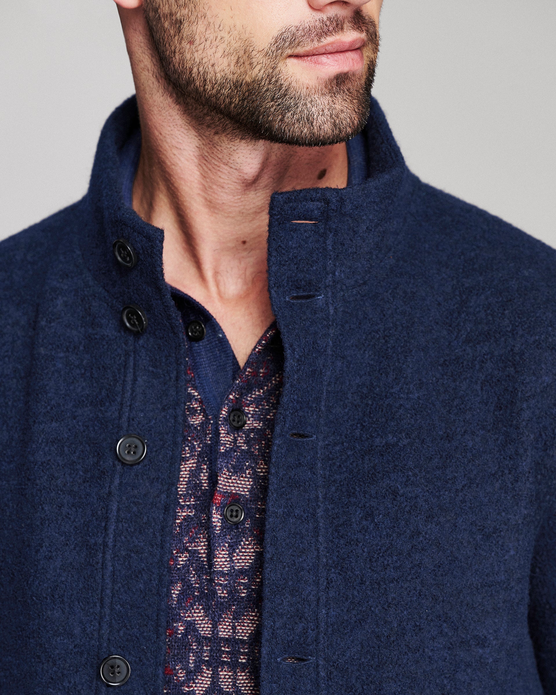 Close-up of a man wearing a navy blue textured wool-blend jacket with a high stand collar and dark buttons, layered over a patterned shirt.
