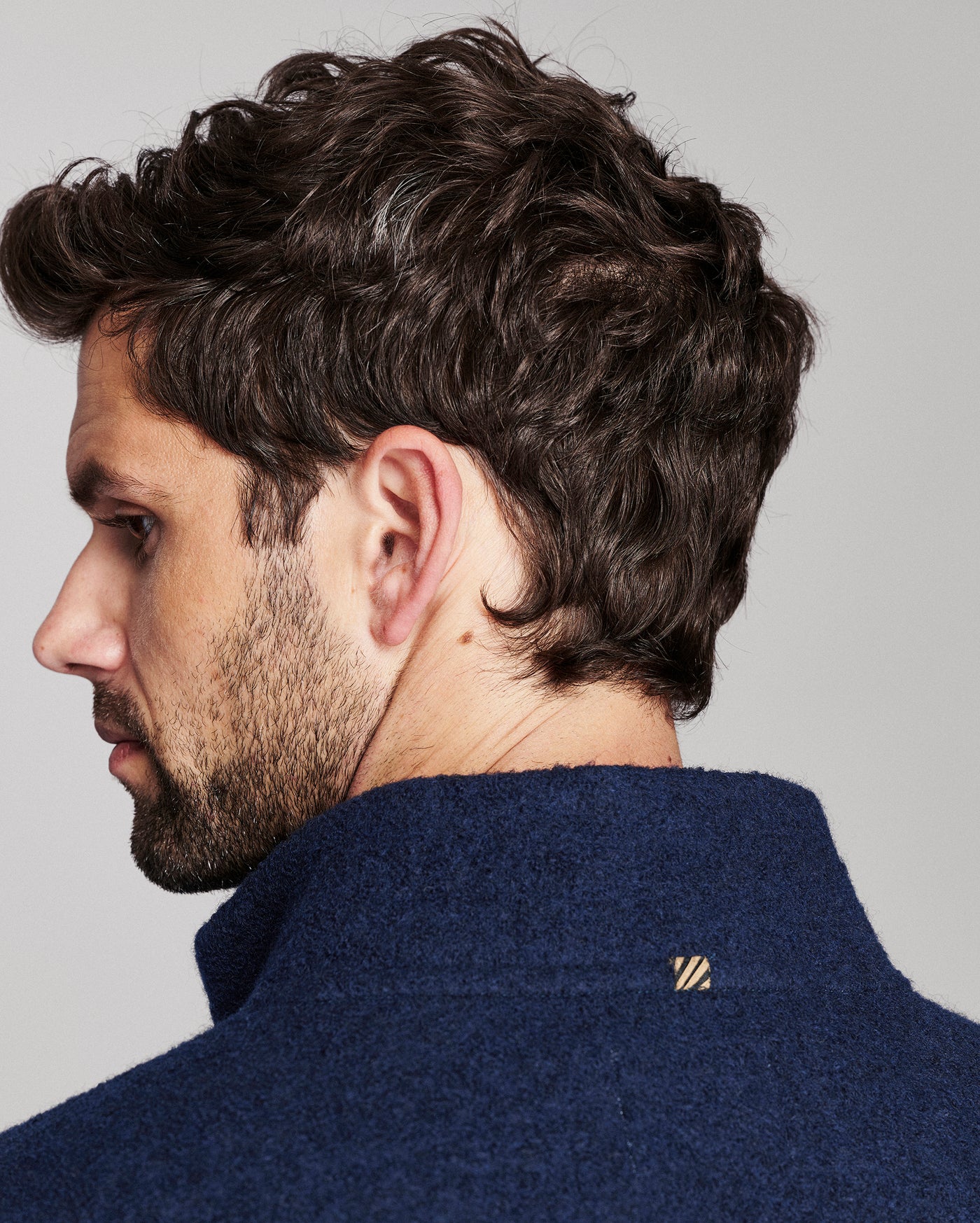 Close-up side profile of a man with dark curly hair wearing a navy blue textured knit sweater, showing a small gold embroidered logo on the collar against a light gray background.