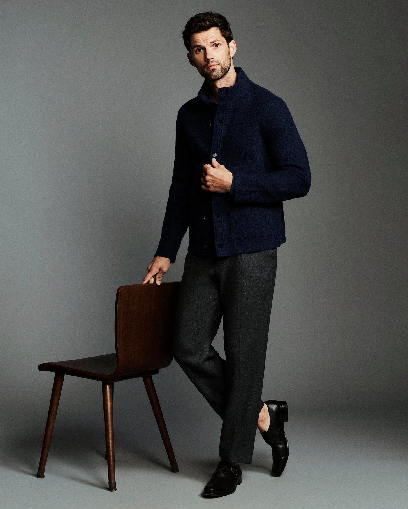 A man modeling a dark navy textured knit cardigan over a collared shirt, paired with charcoal gray trousers and black dress shoes, leaning against a wooden chair against a solid gray studio background.