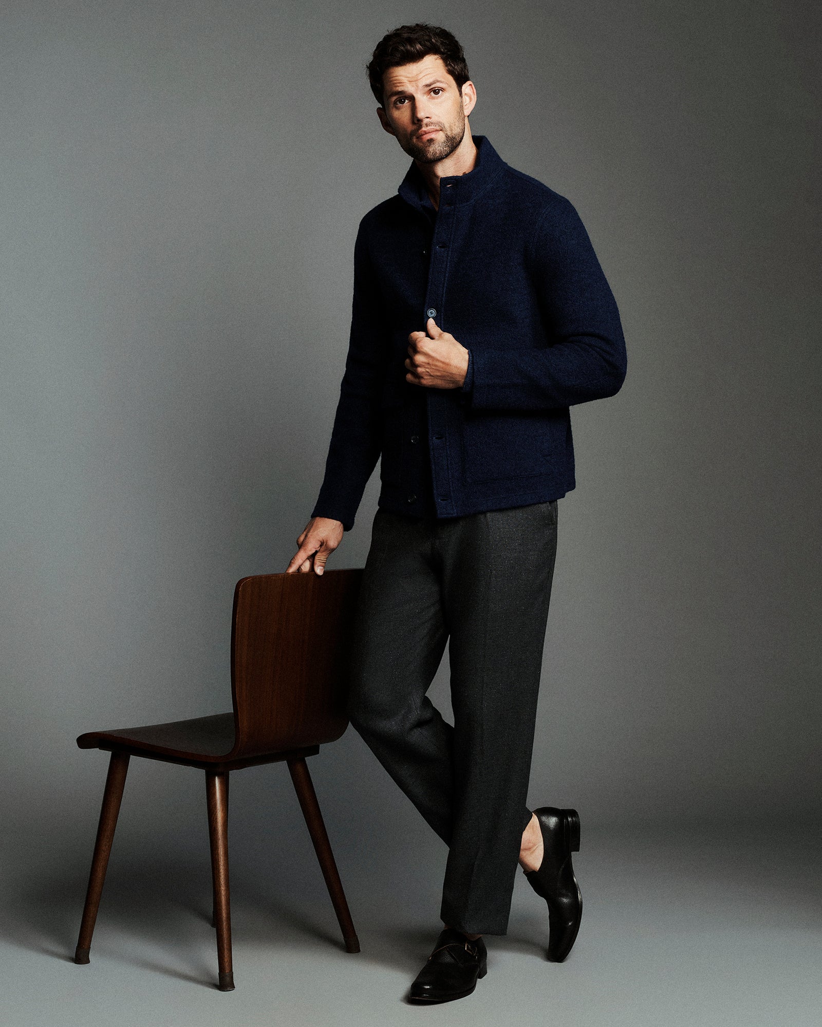 A man modeling a dark navy textured knit cardigan over a collared shirt, paired with charcoal gray trousers and black dress shoes, leaning against a wooden chair against a solid gray studio background.