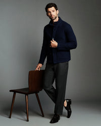 A man modeling a dark navy textured knit cardigan over a collared shirt, paired with charcoal gray trousers and black dress shoes, leaning against a wooden chair against a solid gray studio background.