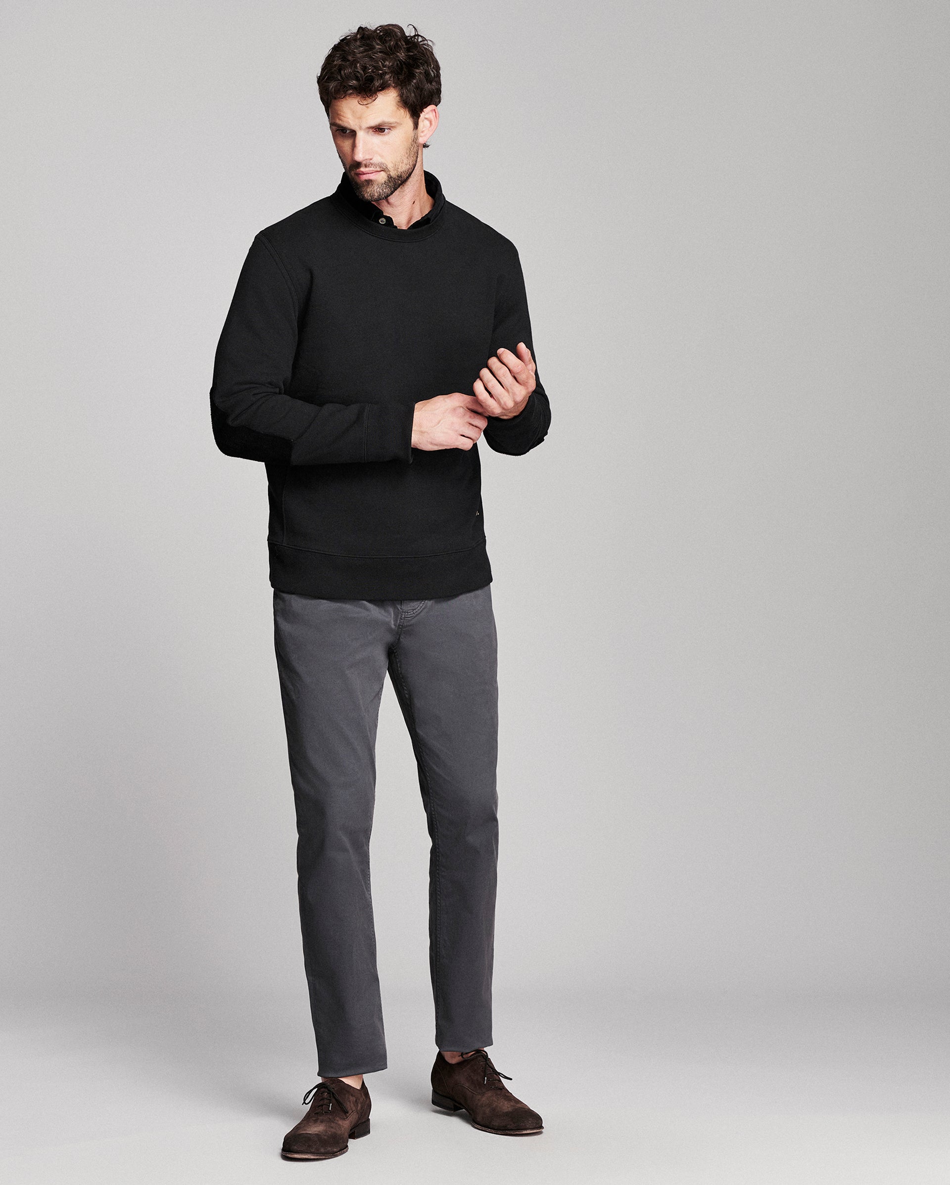 A man modeling a black long-sleeve knit polo-style sweater, paired with gray trousers and dark brown leather shoes, against a light gray studio background.