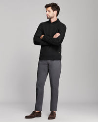 A man wearing a black long-sleeve mock-neck sweater, dark gray trousers, and brown suede loafers, standing with arms crossed against a light gray studio background.