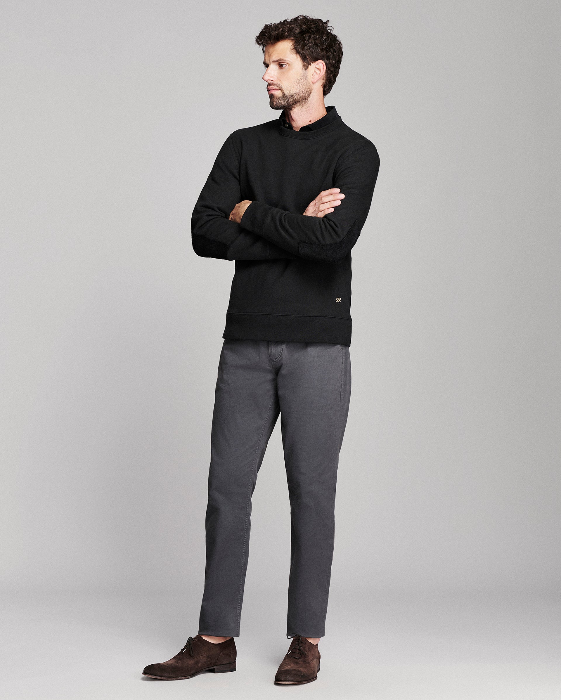 A man wearing a black long-sleeve mock-neck sweater, dark gray trousers, and brown suede loafers, standing with arms crossed against a light gray studio background.