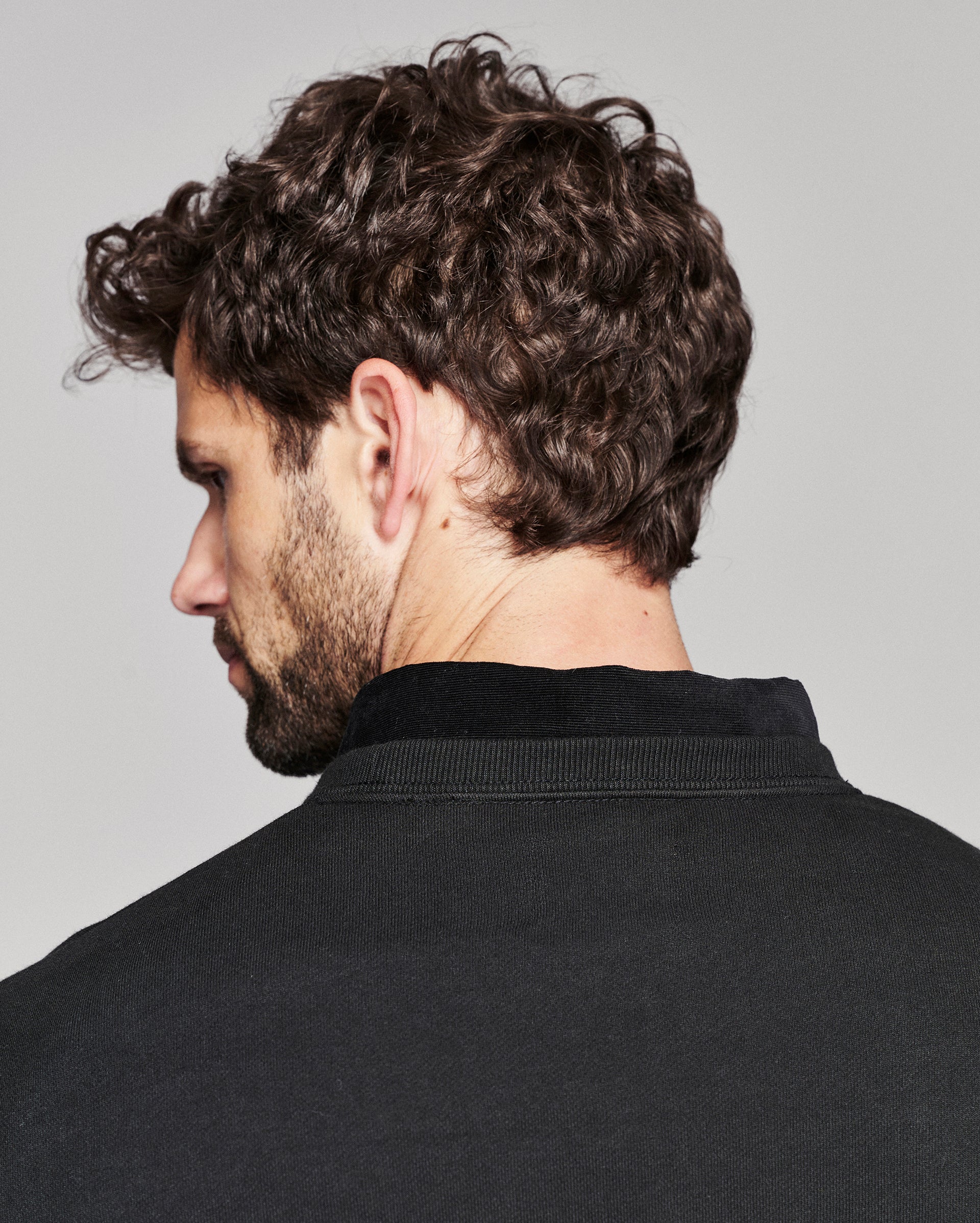 A close-up side profile of a man with curly dark brown hair and a beard, wearing a black crewneck sweater against a light gray background.