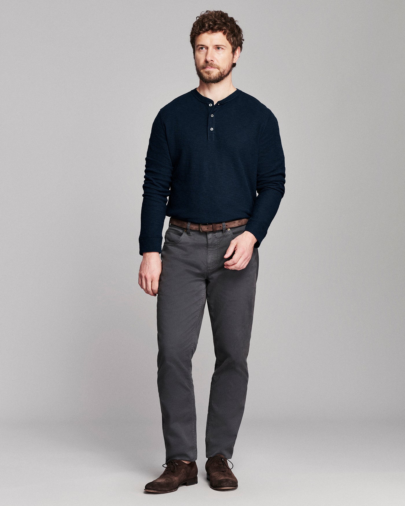A man modeling a dark navy long-sleeve henley shirt, dark gray trousers, a brown belt, and brown leather shoes against a solid light gray studio background.