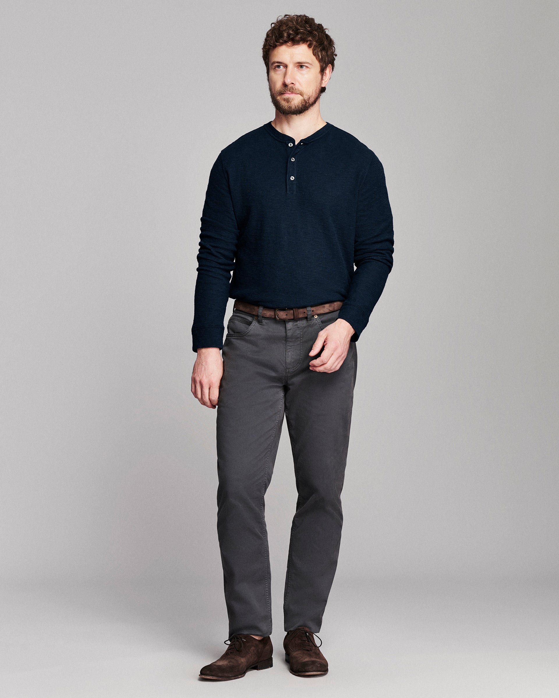 A man modeling a dark navy long-sleeve henley shirt, dark gray trousers, a brown belt, and brown leather shoes against a solid light gray studio background.