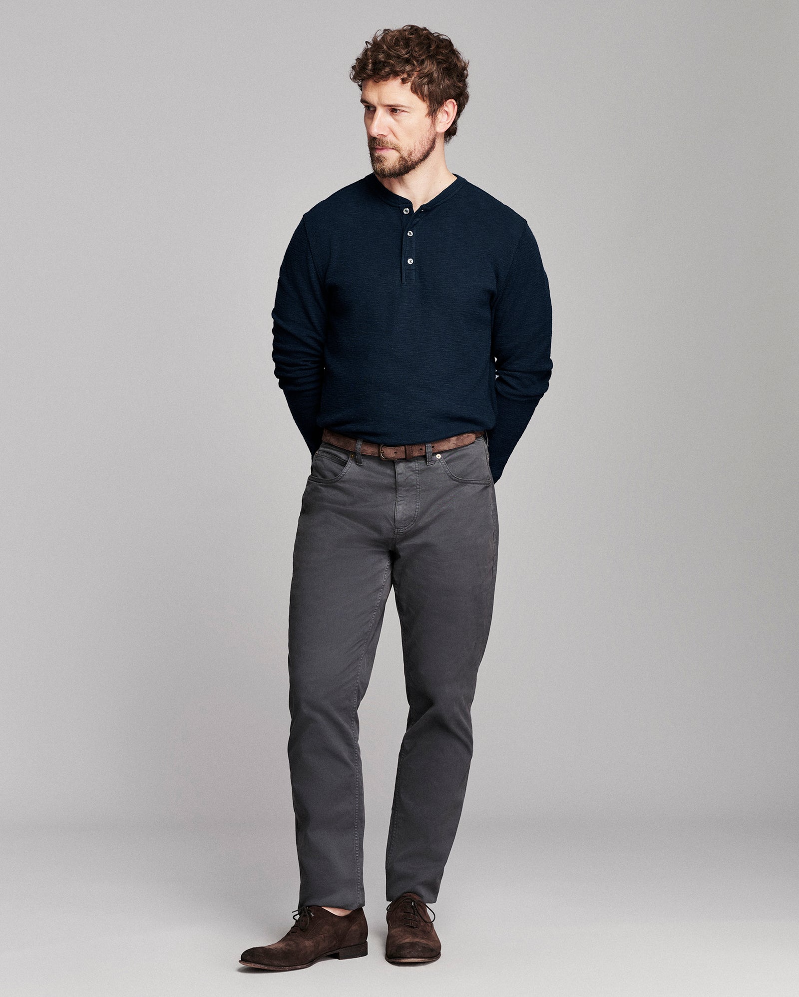 A man modeling a dark navy long-sleeve henley shirt, dark gray trousers, and brown leather shoes against a light gray studio background.