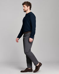 A man in a side profile wearing a dark navy long-sleeve knit sweater, charcoal gray trousers, and dark brown leather shoes, standing against a light gray studio background.