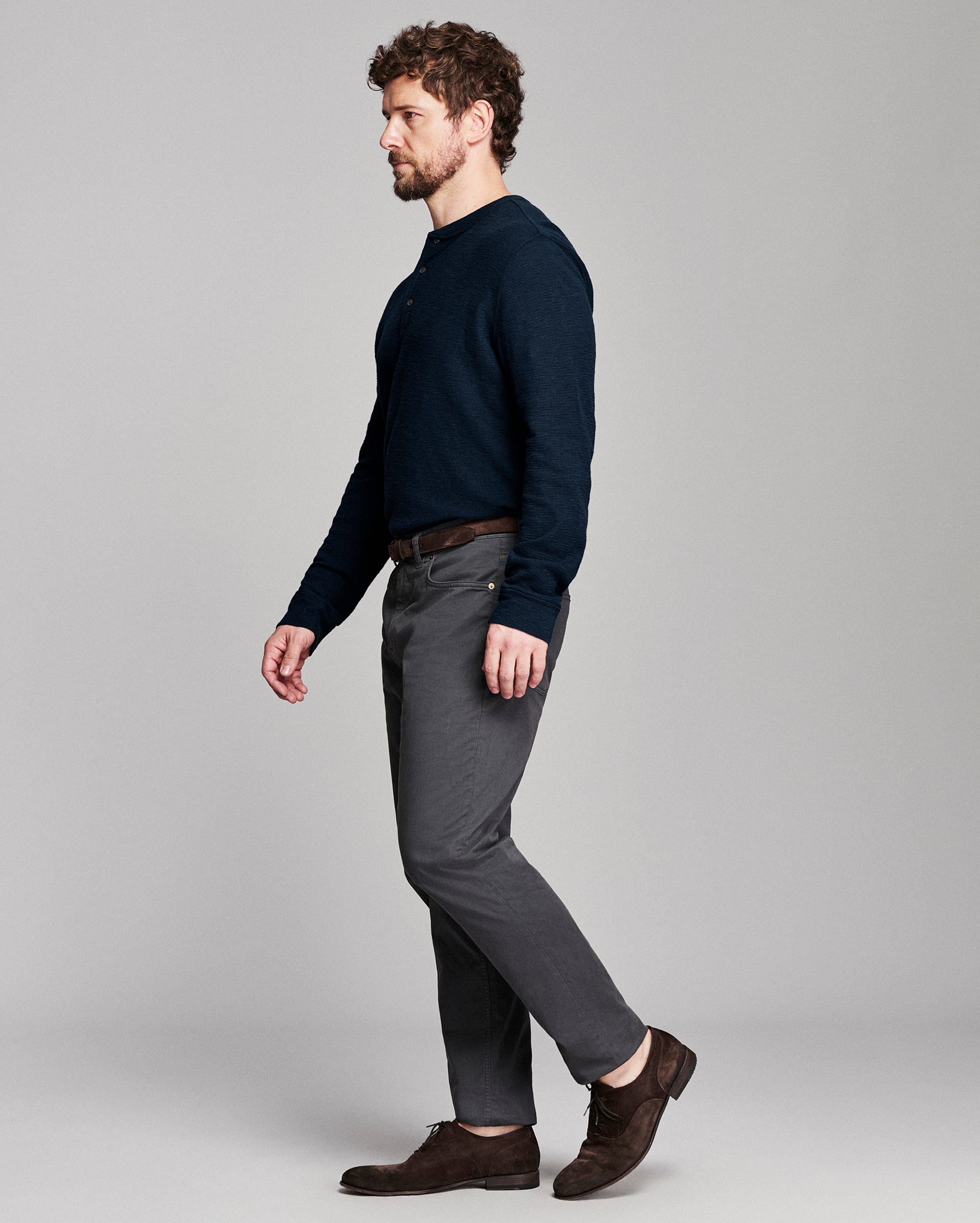 A man in a side profile wearing a dark navy long-sleeve knit sweater, charcoal gray trousers, and dark brown leather shoes, standing against a light gray studio background.