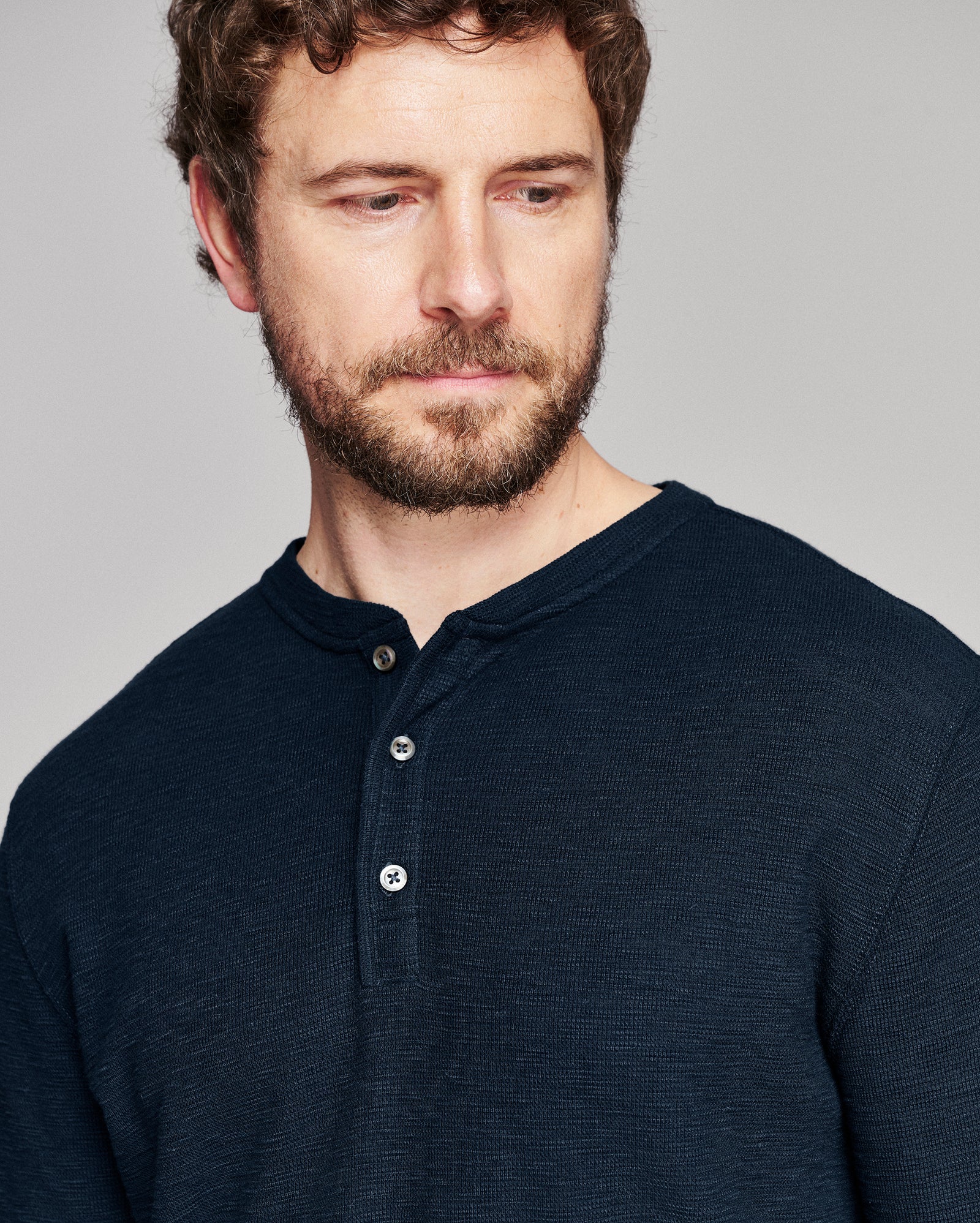 A close-up shot of a man wearing a dark navy blue ribbed henley shirt against a solid gray background.