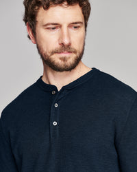 A close-up shot of a man wearing a dark navy blue ribbed henley shirt against a solid gray background.