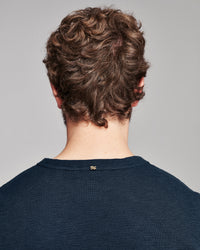 Back view of a person with curly brown hair wearing a dark navy ribbed crewneck sweater against a light gray background.