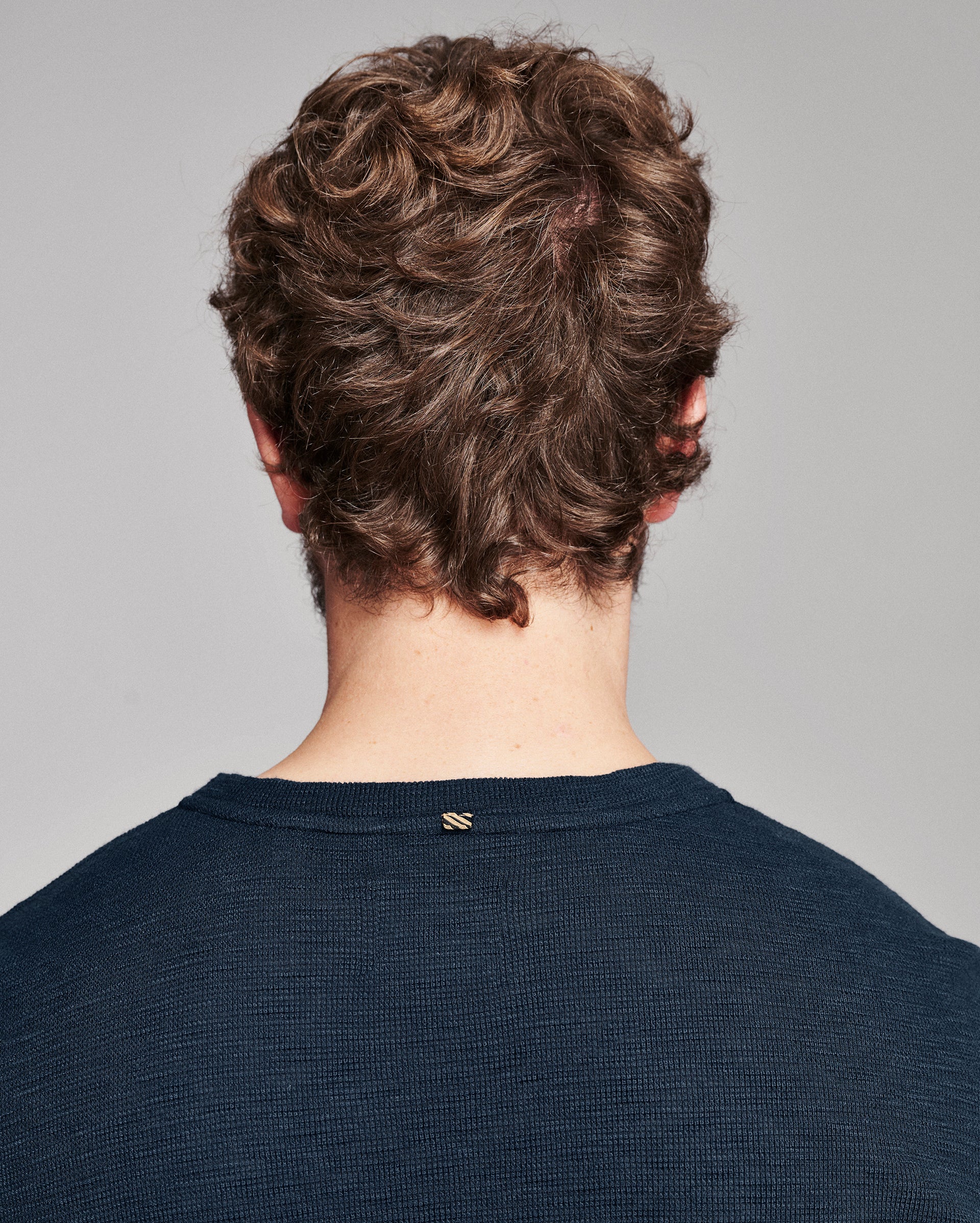 Back view of a person with curly brown hair wearing a dark navy ribbed crewneck sweater against a light gray background.